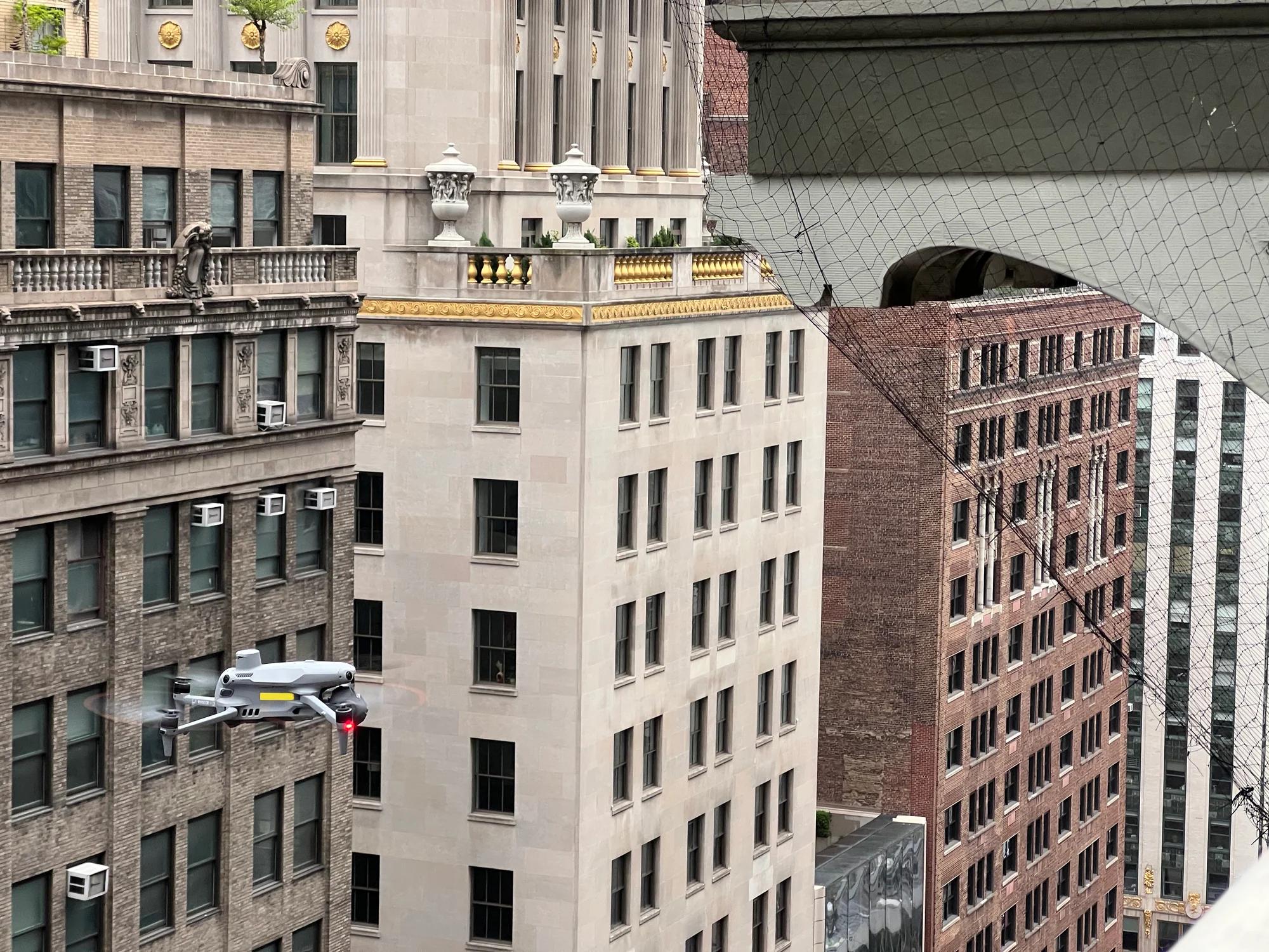 A grey inspection drone hovering between Midtown buildings, the netted cornice at the top right and limestone and brick façades behind