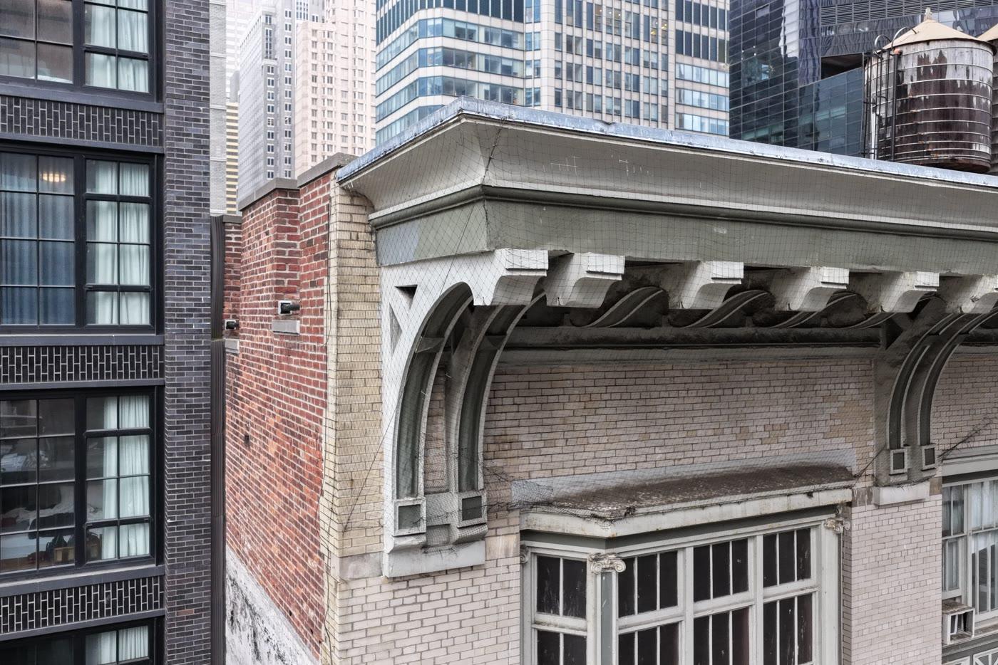 Close-range aerial of a bracketed terracotta cornice under protective netting, with the parapet coping above it
