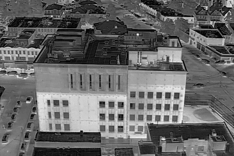 Aerial thermal capture of a masonry building in a dense block, the facade and roof reading pale against the cooler surrounding rooftops and street