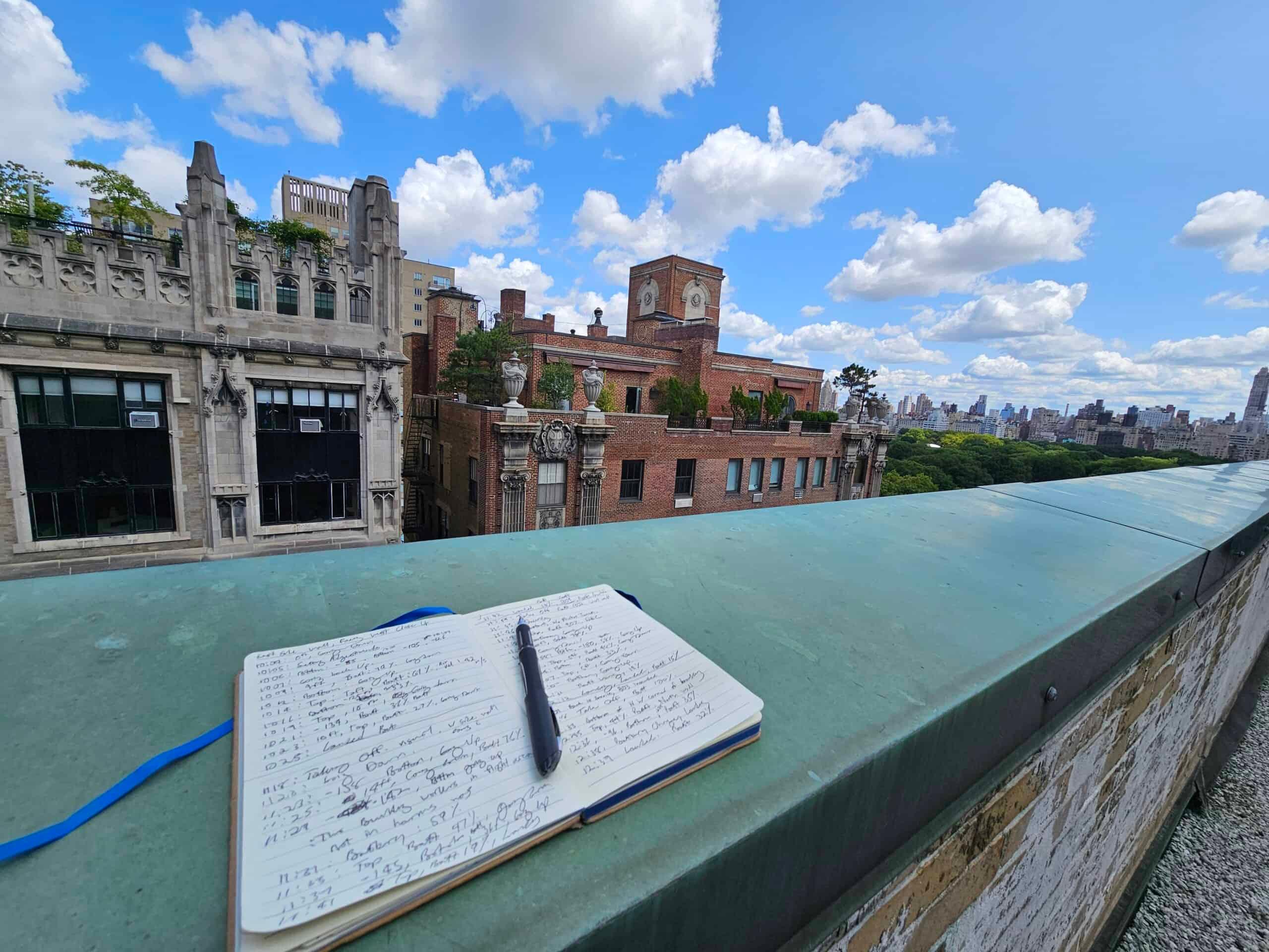 A handwritten flight log open on a copper parapet coping, with Manhattan rooftops and Central Park behind
